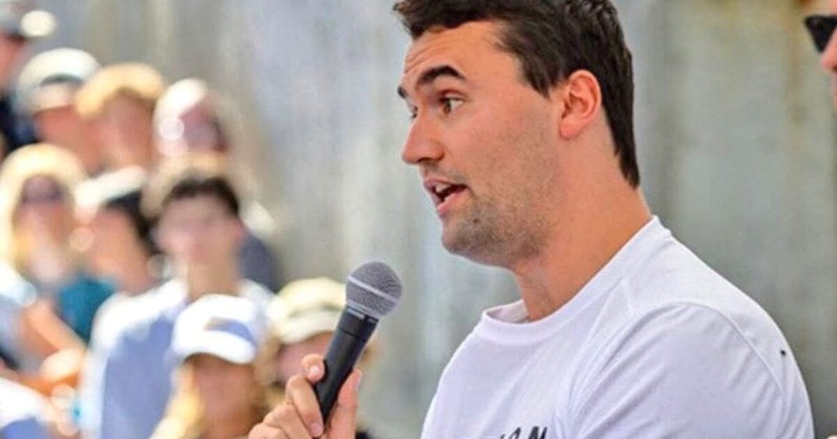 Young man speaking into a microphone at an outdoor event, engaging with an attentive crowd in the background.