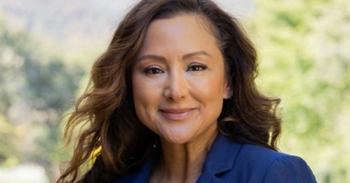 Professional woman with long wavy hair smiling outdoors in a blue blazer against a blurred natural background.