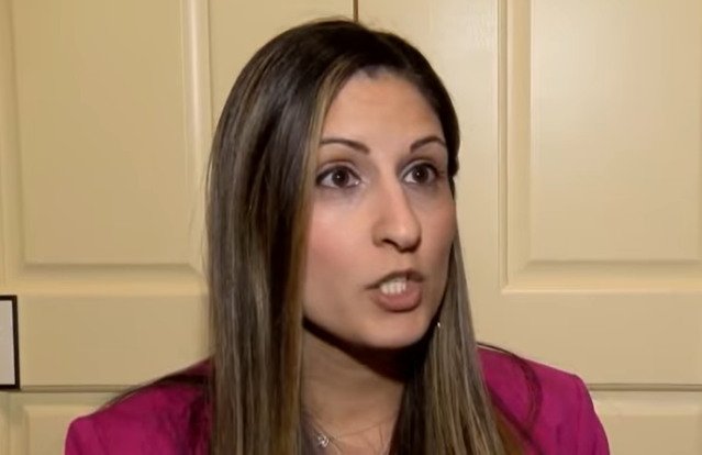 Woman with long hair speaking passionately in a professional setting, wearing a pink blazer against a light-colored background.