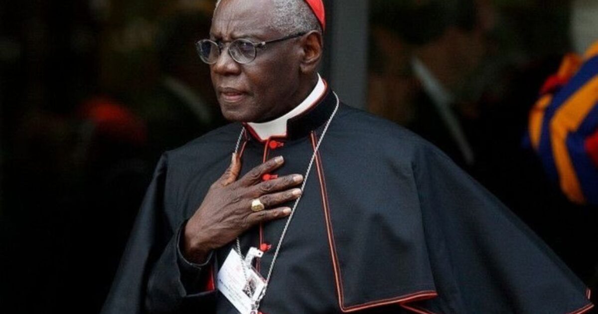 Cardinal dressed in traditional black and red robes, standing outdoors with a thoughtful expression and hand on chest, symbolizing reverence and authority.