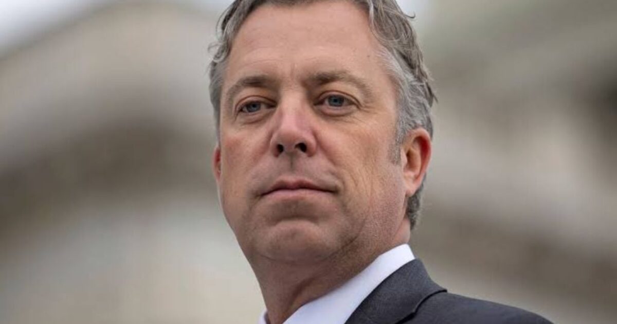 Portrait of a man in a suit with a red tie and American flag pin, standing confidently outdoors.