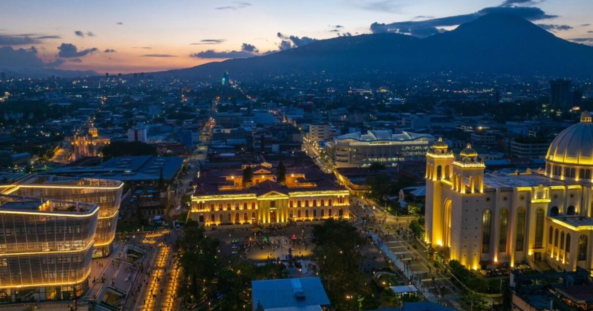 Aerial view of San Salvador at dusk, showcasing illuminated buildings, bustling streets, and the backdrop of a volcanic mountain.