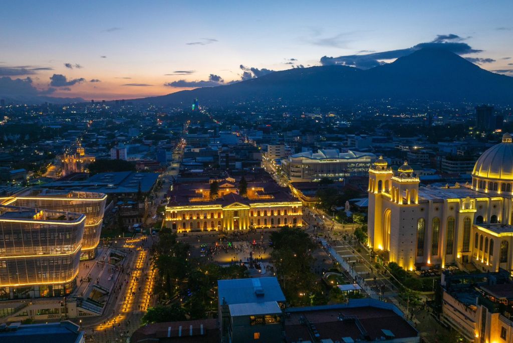 Aerial view of San Salvador at dusk, showcasing illuminated buildings, bustling streets, and the backdrop of a volcanic mountain.