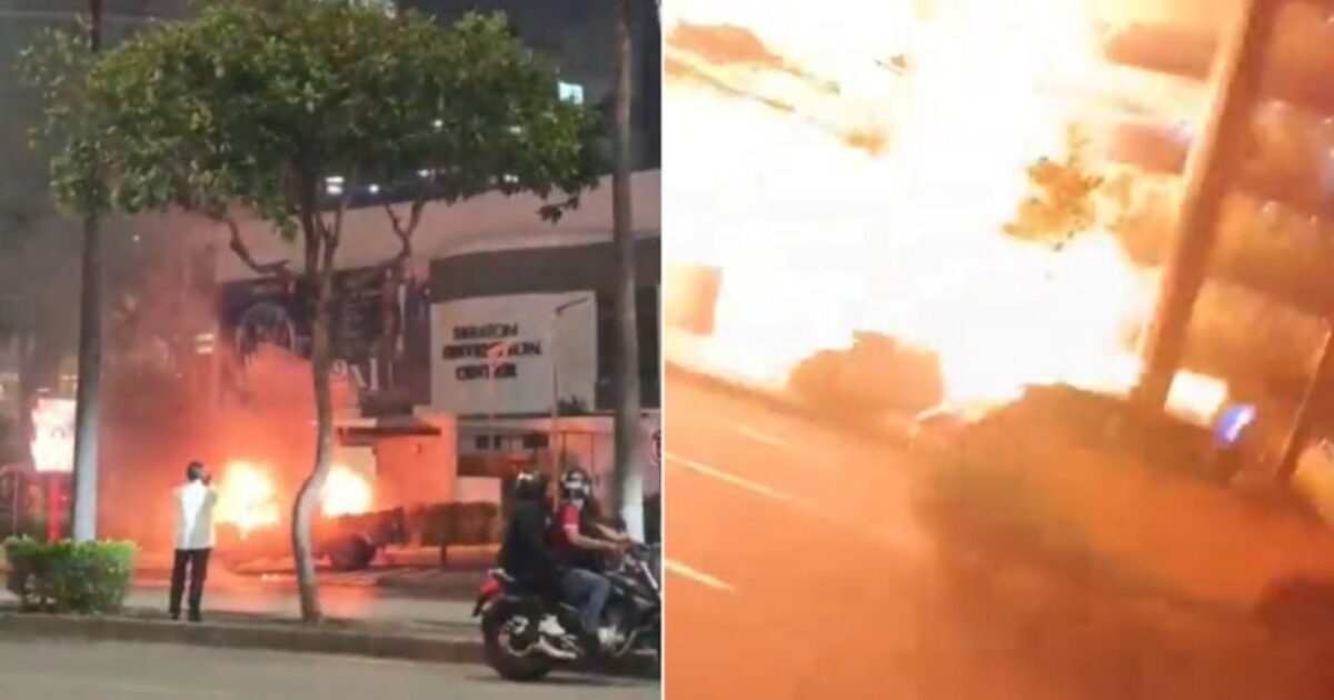 Fire erupts in an urban area, with onlookers observing the blaze near buildings and vehicles at night.