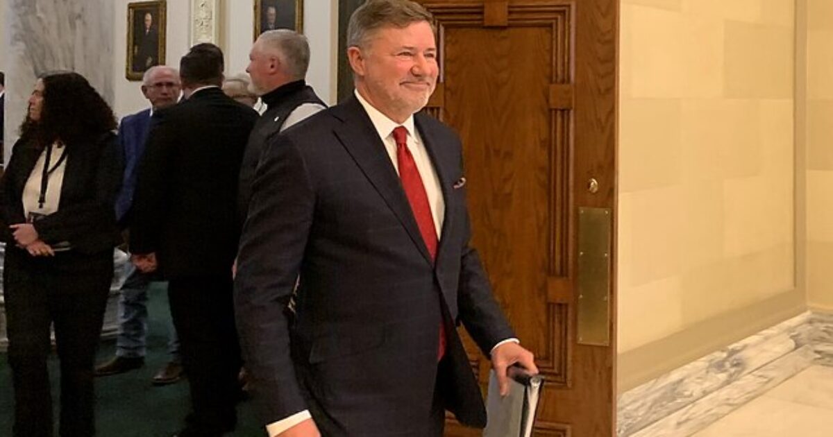 Smiling man in a suit entering a building with historical decor, surrounded by people in formal attire.