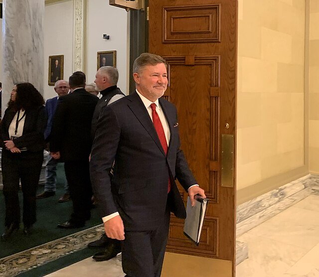 Smiling man in a suit entering a building with historical decor, surrounded by people in formal attire.