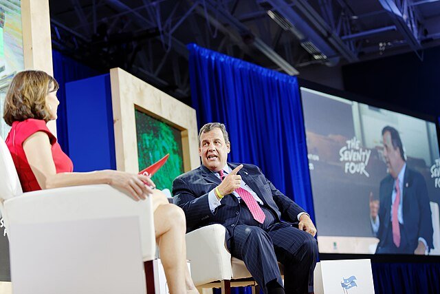 Panel discussion featuring a man and a woman engaged in conversation on stage, with a large screen displaying the event title in the background.