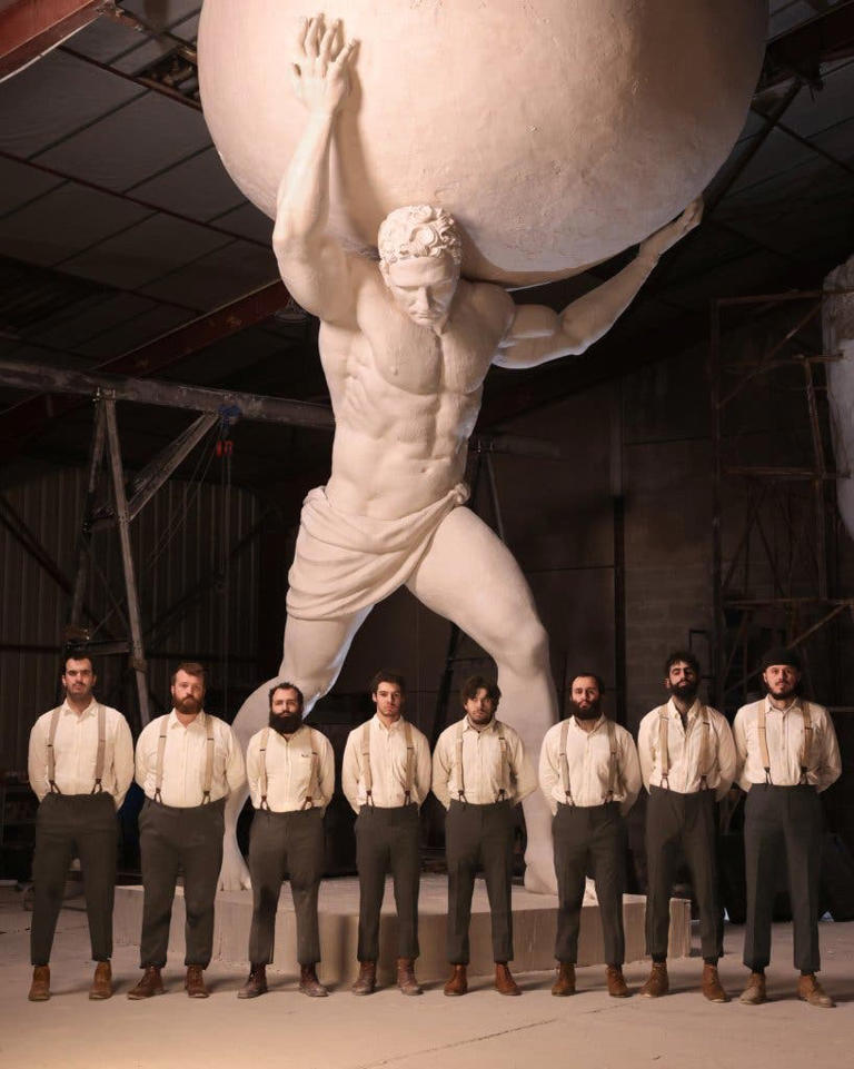Group of men in vintage attire standing in front of a large statue of Atlas holding a globe in a workshop setting.