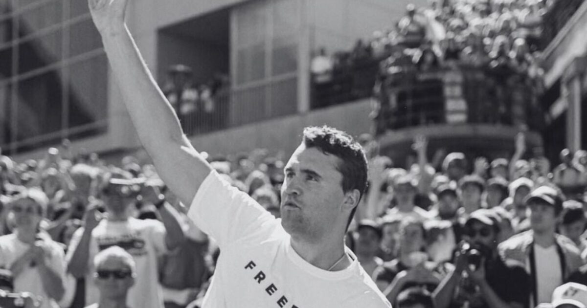 Man in a white t-shirt with "FREEDOM" raises his hand in celebration, surrounded by a cheering crowd at an outdoor event.