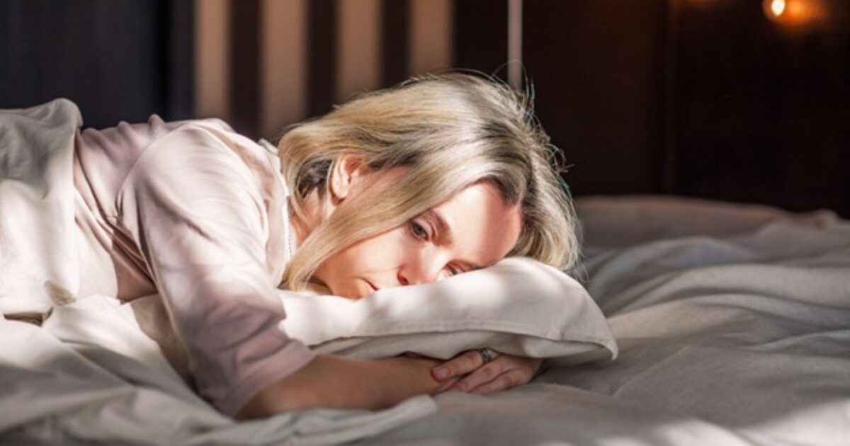 Woman lying on a bed with a contemplative expression, surrounded by soft bedding and warm light, conveying a sense of introspection and tranquility.