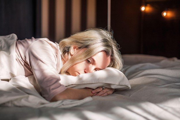Woman lying on a bed with a contemplative expression, surrounded by soft bedding and warm light, conveying a sense of introspection and tranquility.