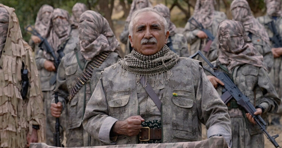 A leader addresses armed militants in camouflage uniforms, emphasizing unity and strength in a mountainous outdoor setting.