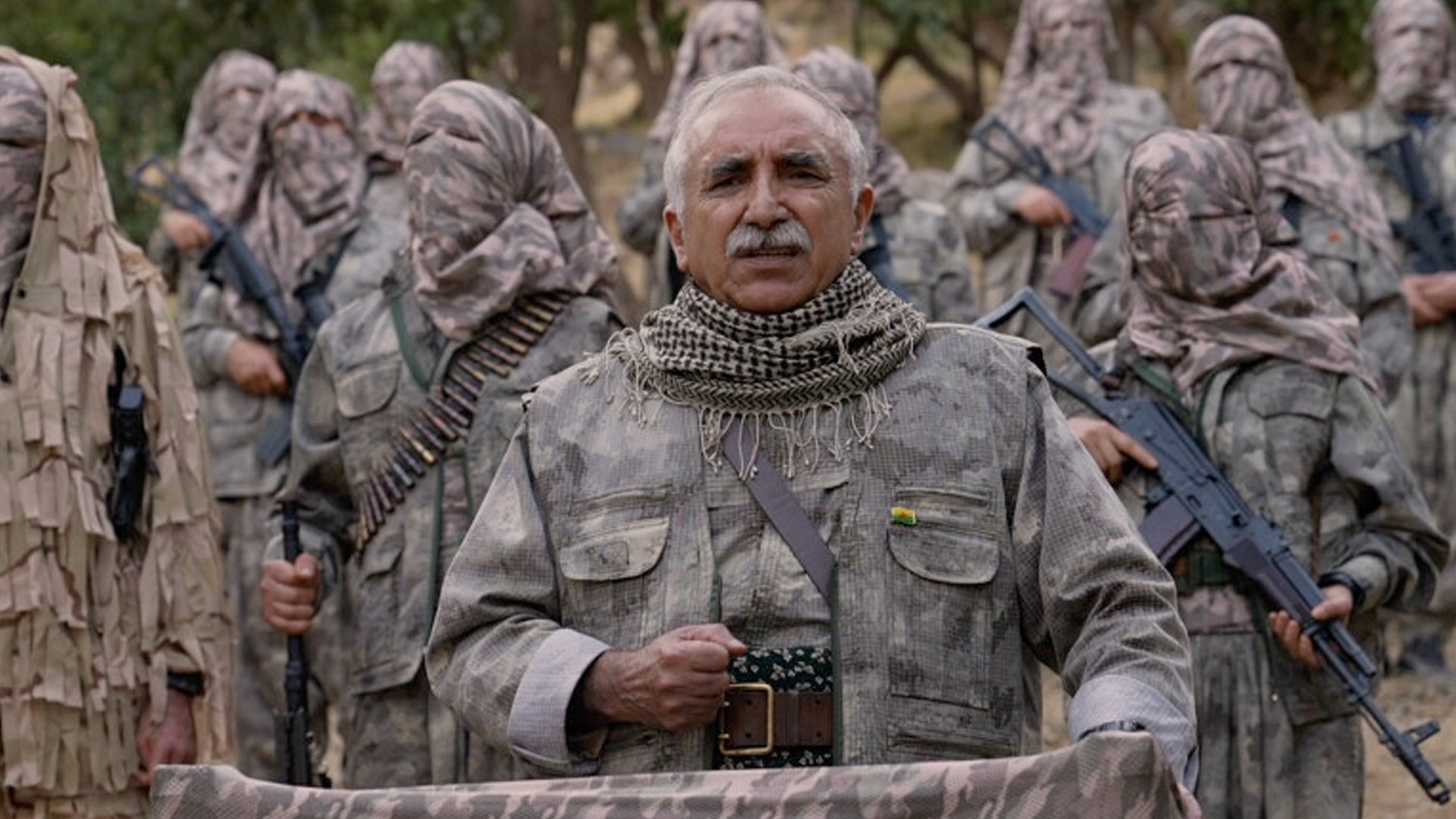 A leader addresses armed militants in camouflage uniforms, emphasizing unity and strength in a mountainous outdoor setting.