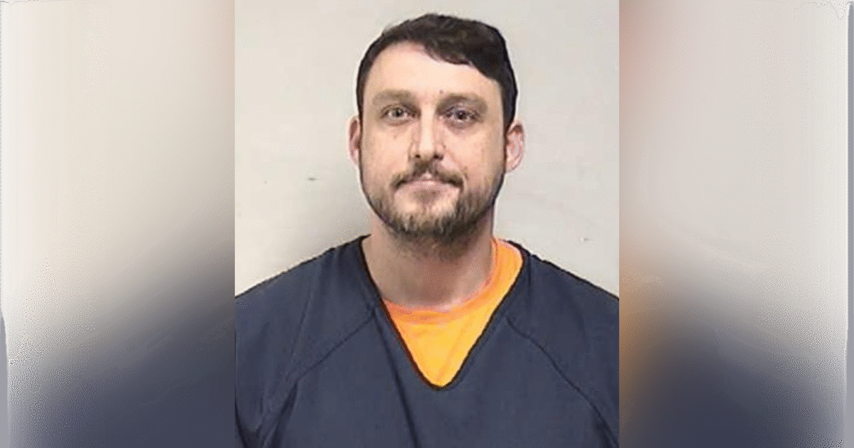 Mugshot of a male inmate wearing a blue jumpsuit and orange shirt, posing for an official identification photo against a plain background.