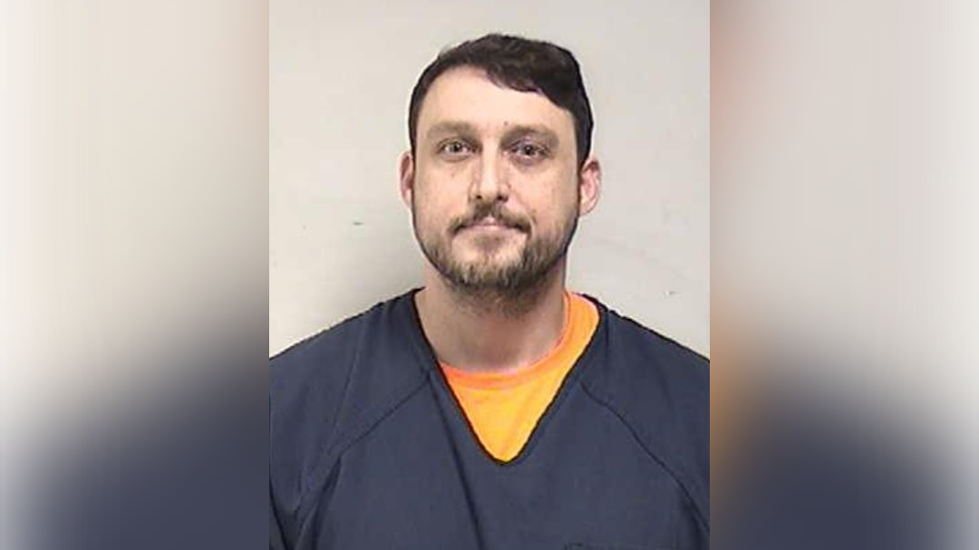 Mugshot of a male inmate wearing a blue jumpsuit and orange shirt, posing for an official identification photo against a plain background.