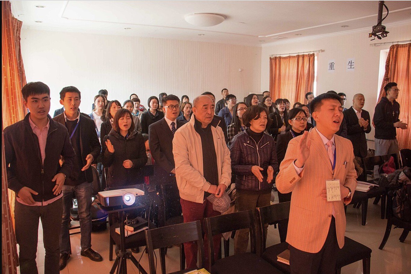A diverse group of people engaged in a worship service, singing and expressing devotion in a bright, indoor setting.
