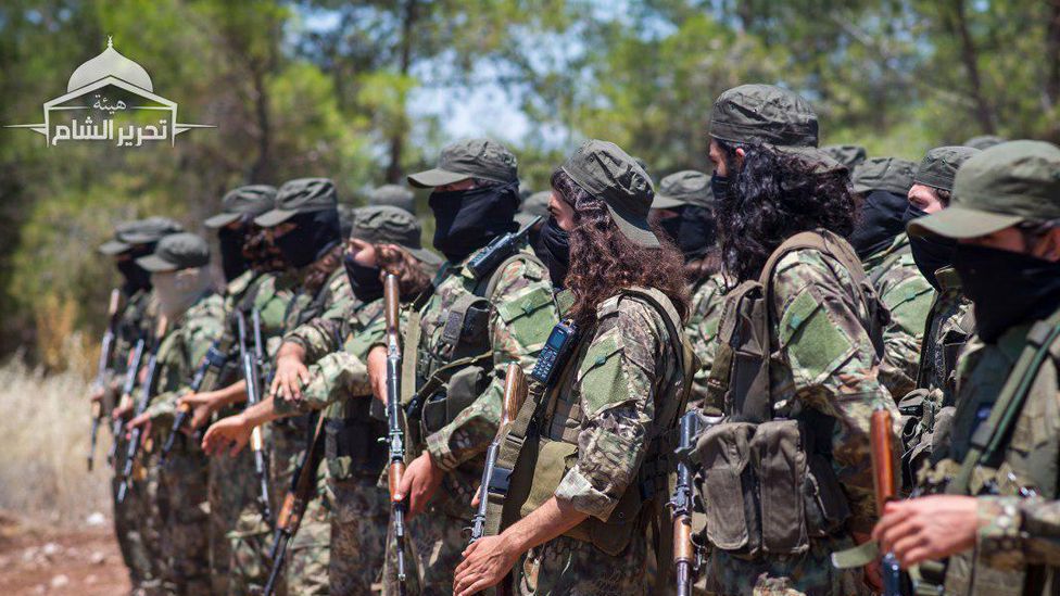 Masked fighters in military attire stand in formation, holding weapons, during a training exercise in a wooded area.