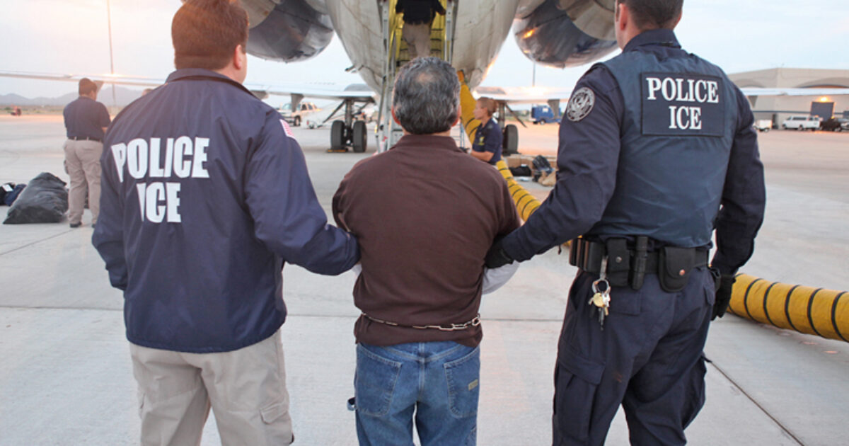 Police officers from ICE escorting a detained individual toward an aircraft at an airport, highlighting immigration enforcement activities.