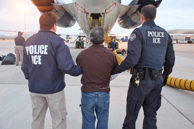 Police officers from ICE escorting a detained individual toward an aircraft at an airport, highlighting immigration enforcement activities.