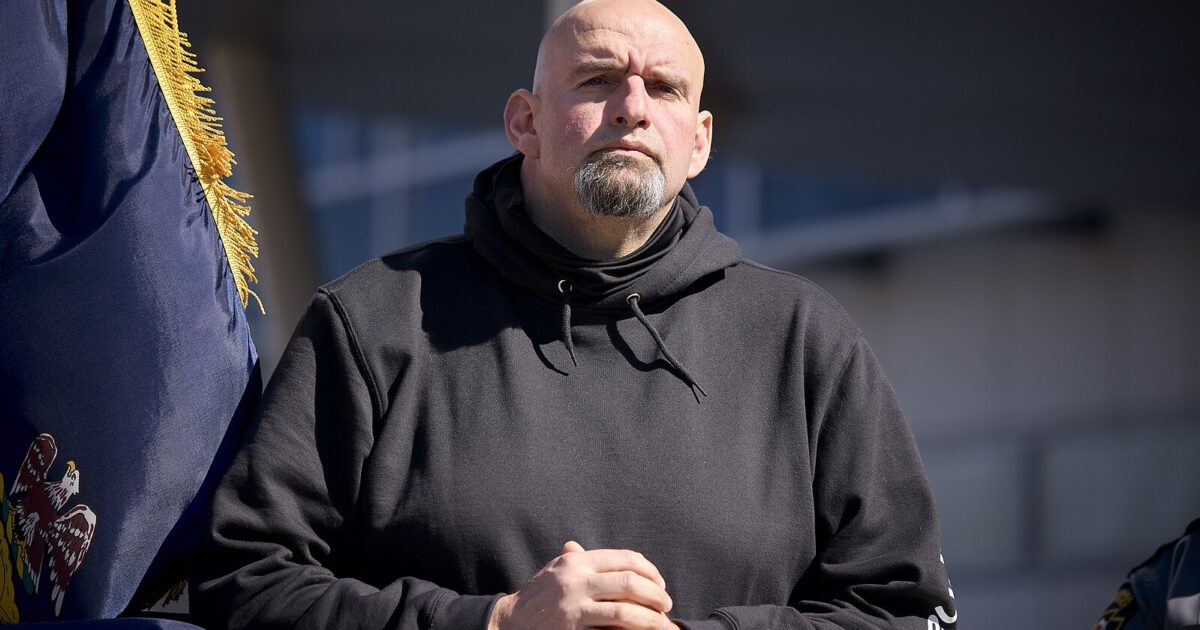 Man in a black hoodie stands confidently with hands clasped, framed by a flag, during an outdoor event in bright sunlight.
