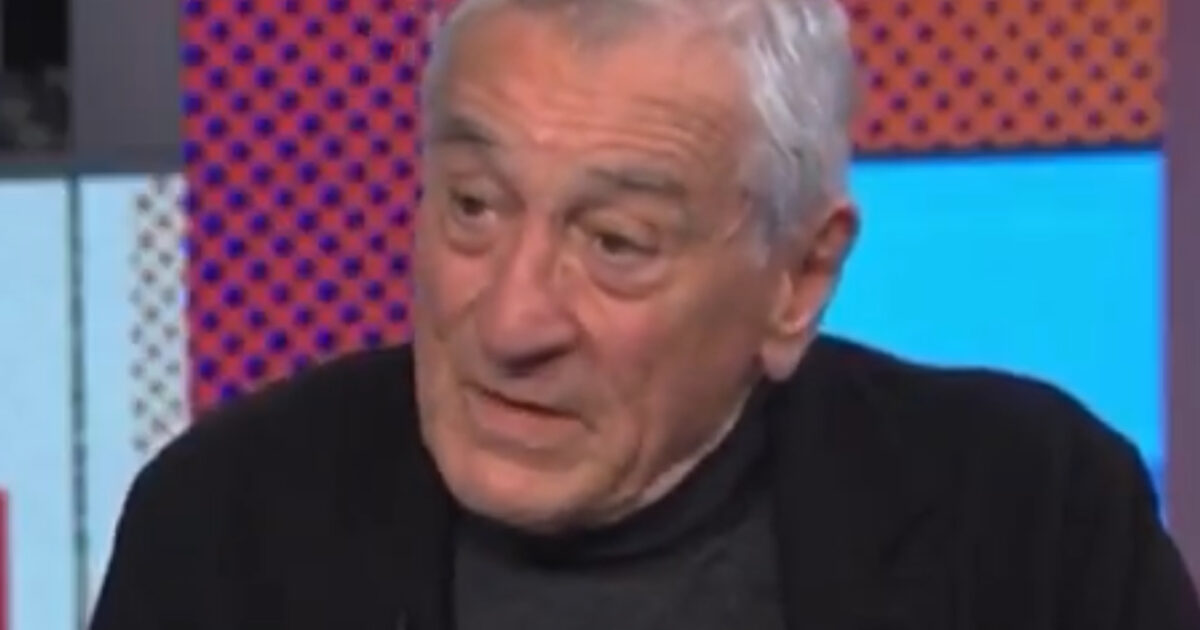 An older man with gray hair speaks thoughtfully during an interview, dressed in a black jacket against a colorful background.