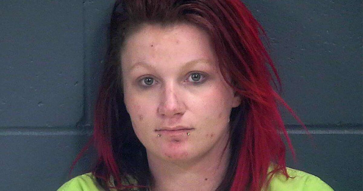 Mugshot of a woman with red and black hair wearing a green shirt, against a gray wall, displaying facial piercings and visible skin blemishes.