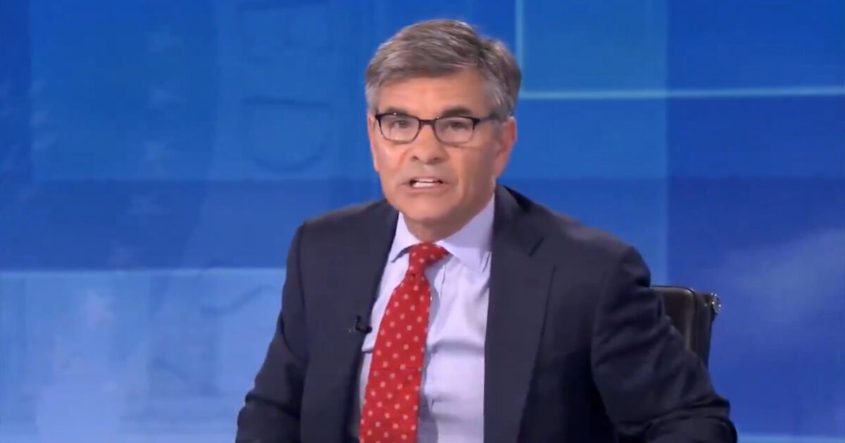 News anchor presenting a segment, wearing glasses and a suit with a red tie, against a blue background in a studio setting.