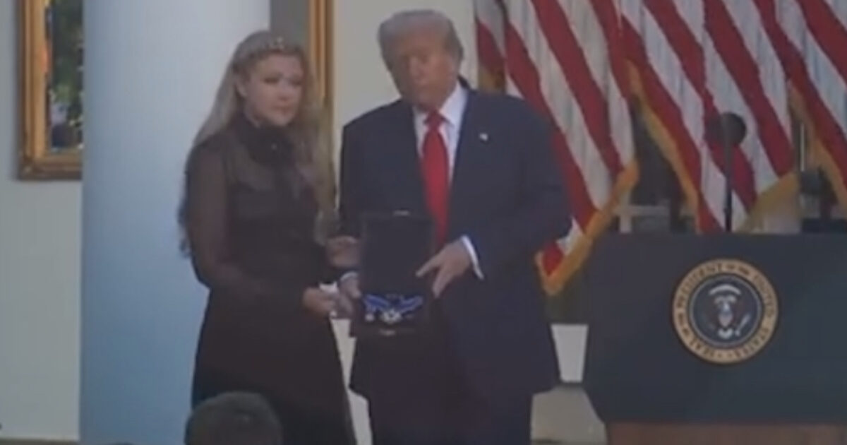 Donald Trump presents an award to a woman during a ceremony in front of American flags in a formal setting.