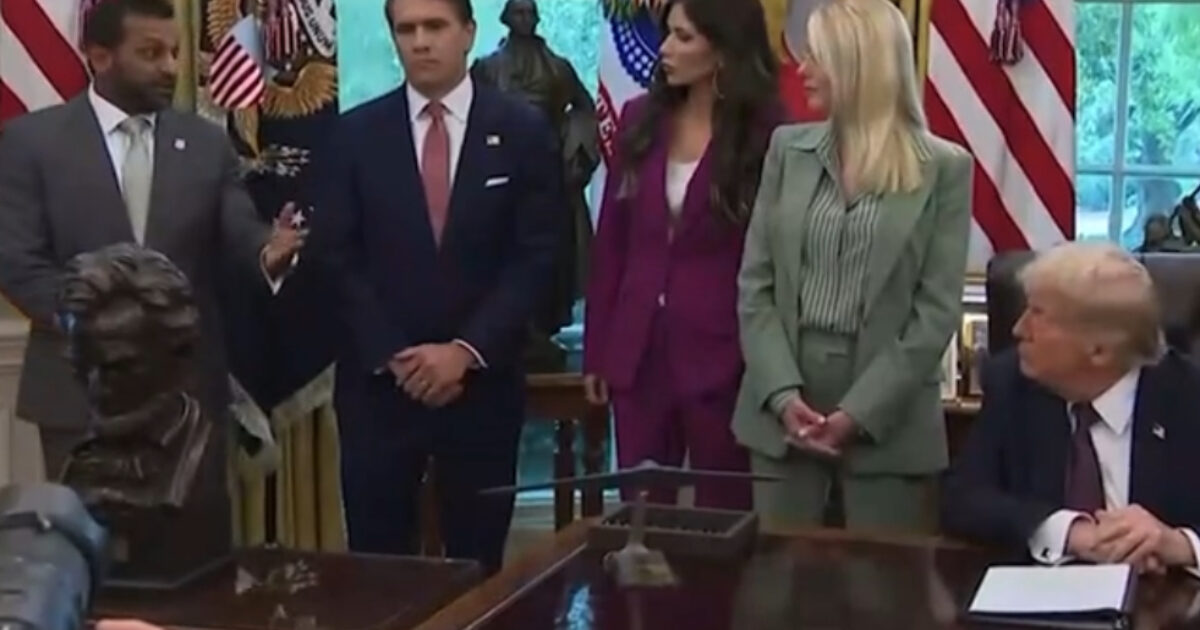 Group of officials speaking in the Oval Office with Trump, featuring flags and a bust in the foreground.