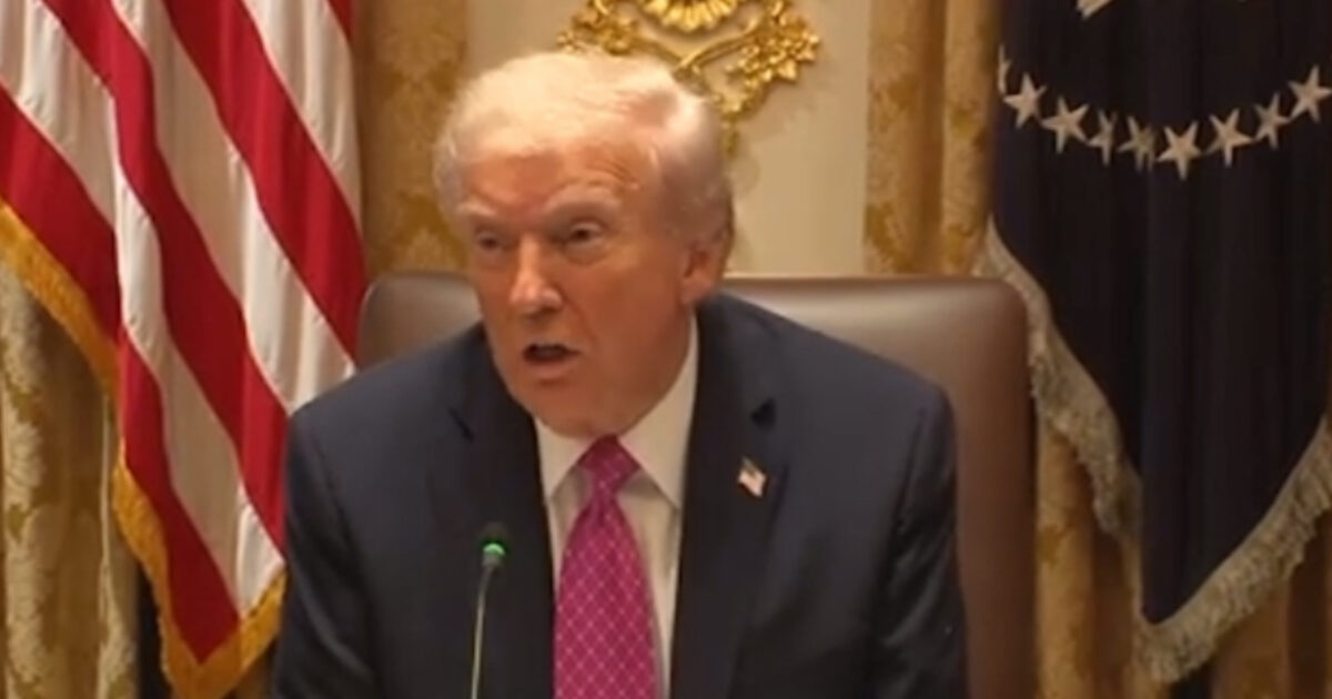 Donald Trump speaking during a meeting, seated at a table with U.S. flags and presidential decor in the background.
