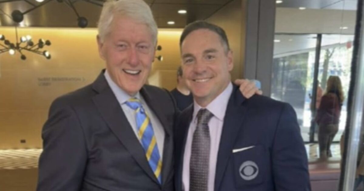 Former President Bill Clinton poses with a man in a suit, both smiling, in a modern office setting with large windows.
