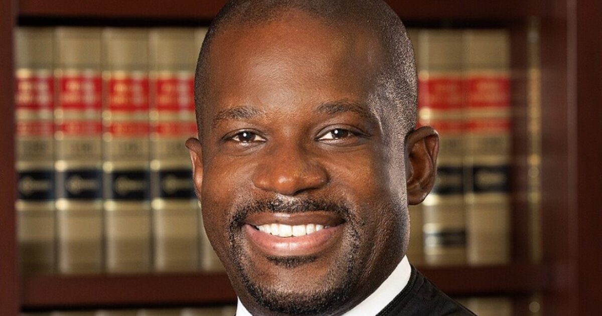 Portrait of a smiling judge in a formal robe, standing in front of legal books in a professional setting.