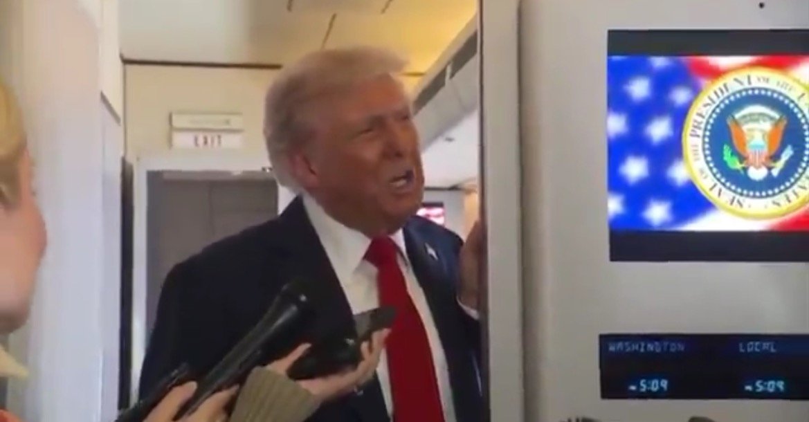 Donald Trump speaking to reporters on an airplane, with a presidential seal displayed on a screen in the background.