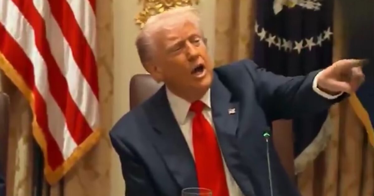 Donald Trump gesturing while speaking at a formal meeting, with the American flag in the background.