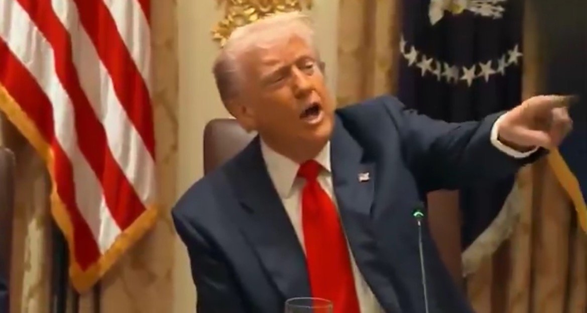 Donald Trump gesturing while speaking at a formal meeting, with the American flag in the background.