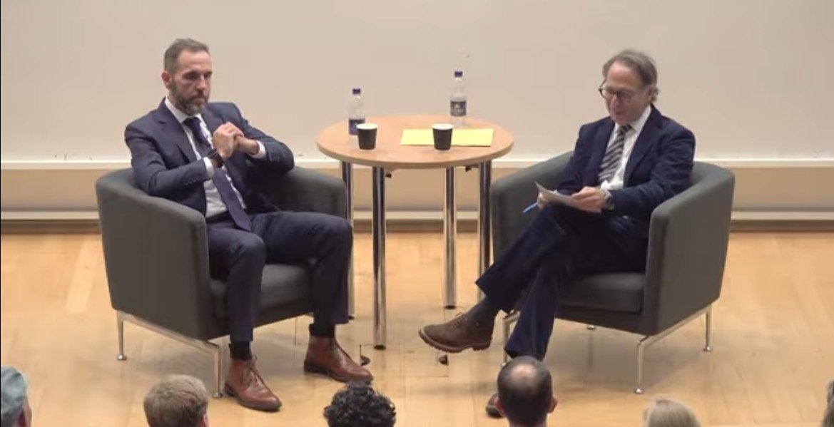 Two men seated in armchairs during an engaging discussion, with a table featuring drinks and notes in a lecture hall setting.