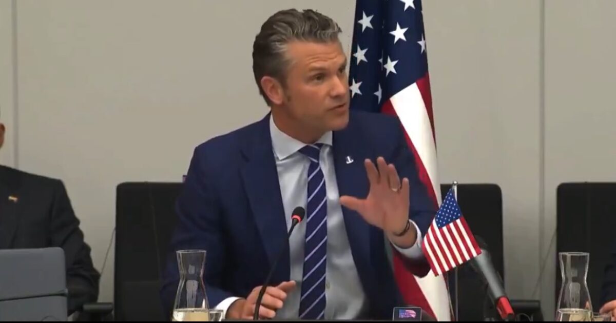 Man in a suit speaking during a formal meeting, with a U.S. flag in the background and a glass of water on the table.