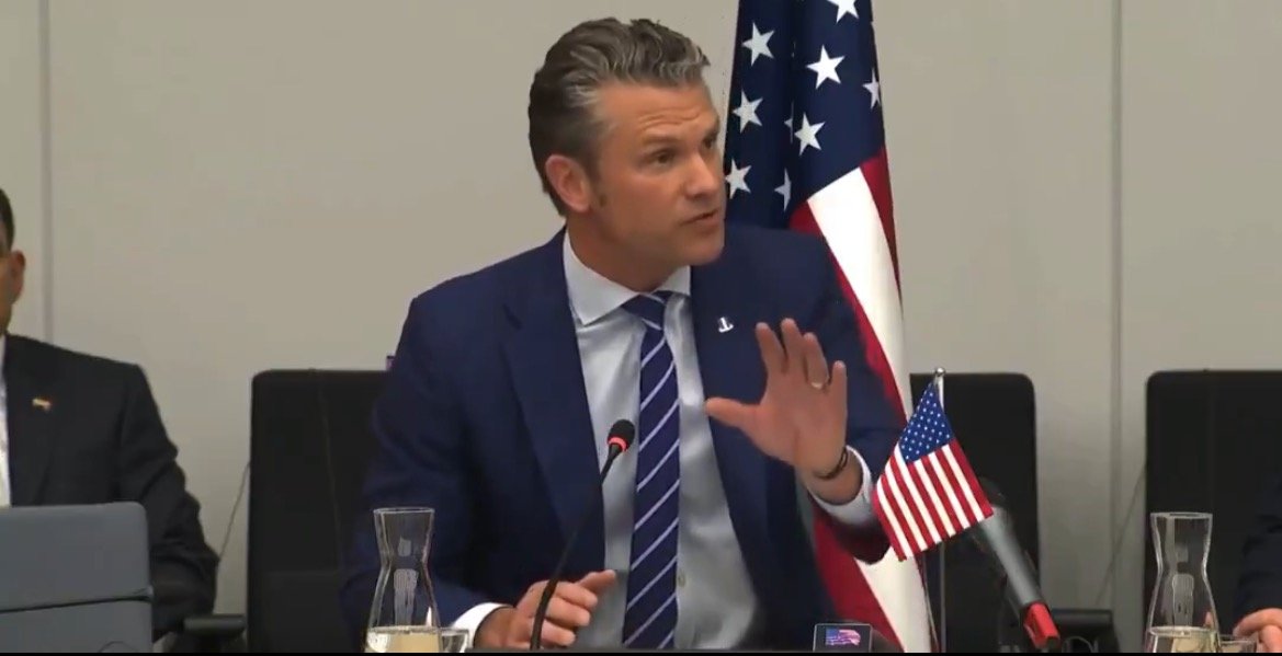 Man in a suit speaking during a formal meeting, with a U.S. flag in the background and a glass of water on the table.