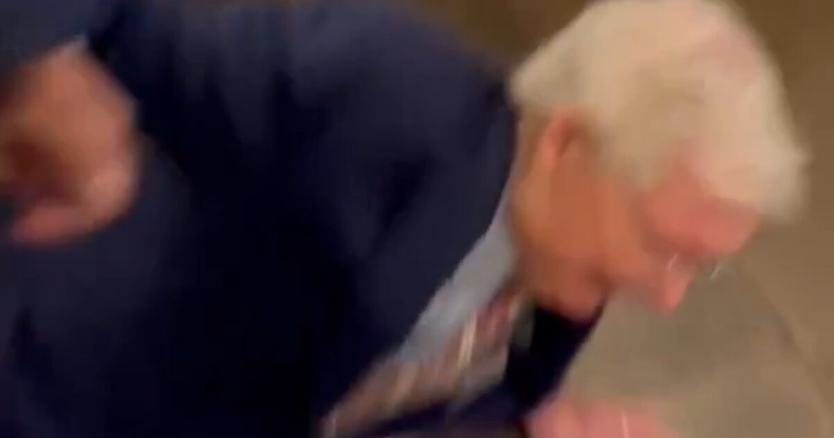 A man in a suit is seen falling to the ground in a blurred indoor setting, capturing a moment of unexpected motion.