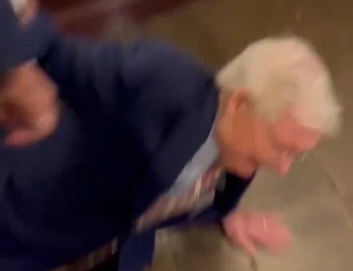 A man in a suit is seen falling to the ground in a blurred indoor setting, capturing a moment of unexpected motion.