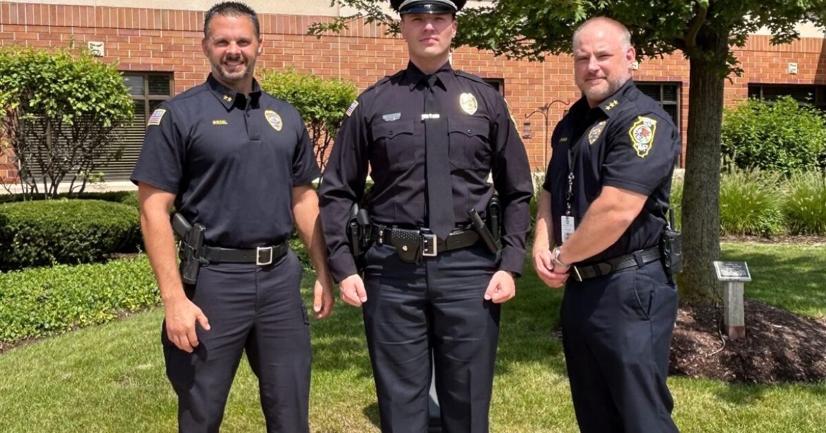 Three police officers in uniform pose outdoors in front of a brick building surrounded by greenery, showcasing community law enforcement.