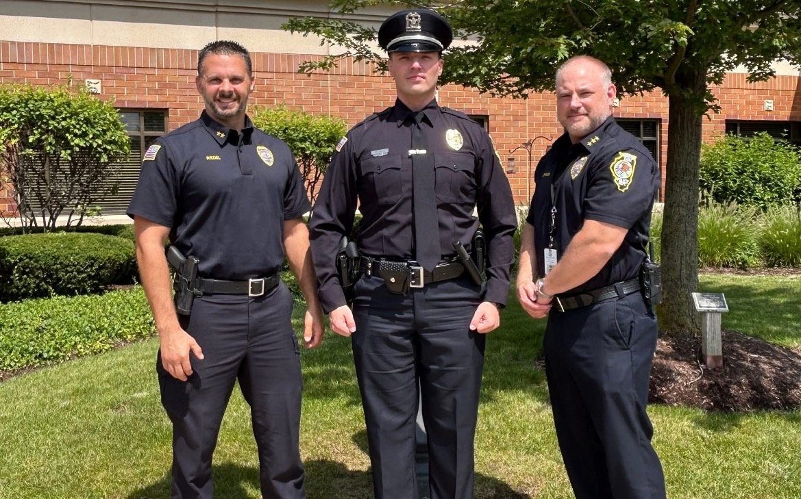 Three police officers in uniform pose outdoors in front of a brick building surrounded by greenery, showcasing community law enforcement.