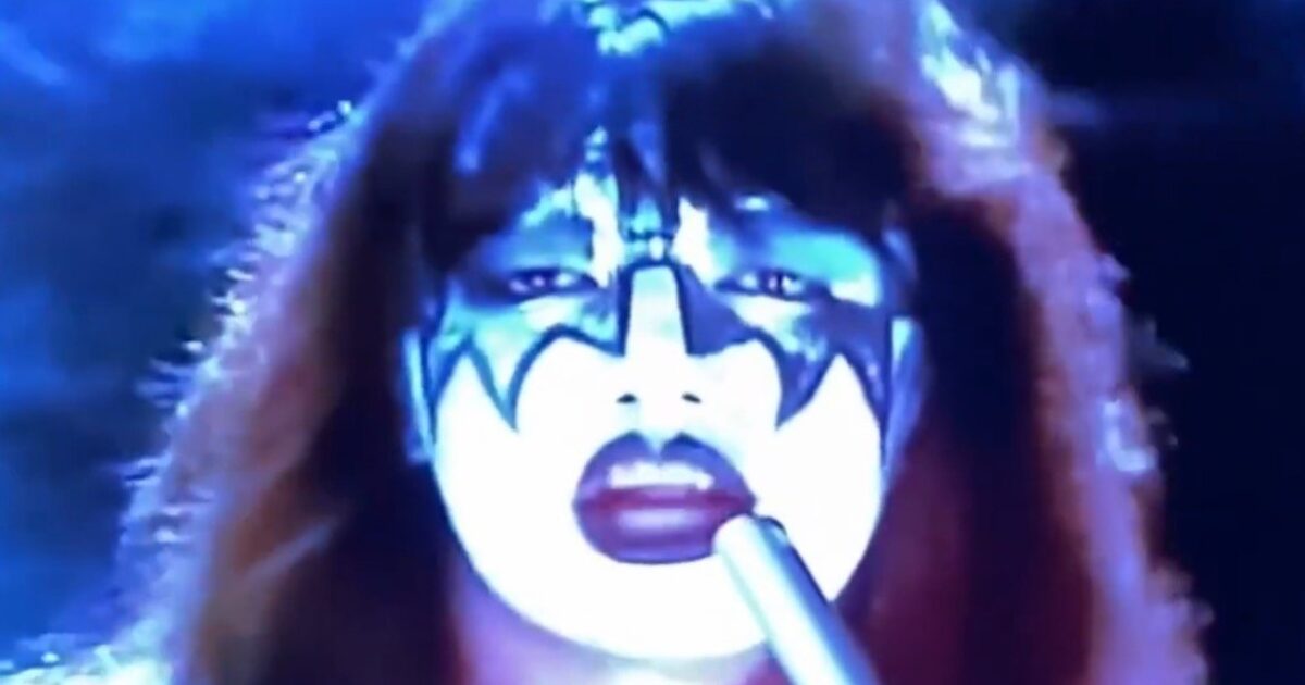 Close-up of a performer with dramatic face paint and long hair, set against a blue background, holding a microphone in a dynamic pose.