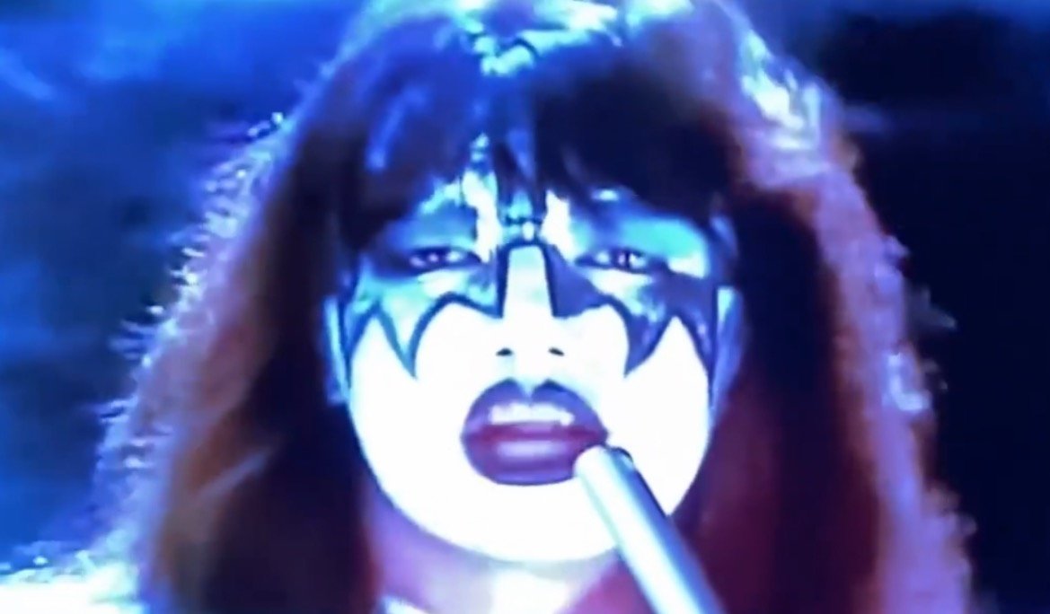 Close-up of a performer with dramatic face paint and long hair, set against a blue background, holding a microphone in a dynamic pose.