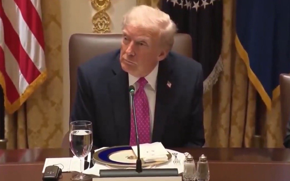 Donald Trump seated at a formal dining table with a glass of water and a plate, during an official meeting.
