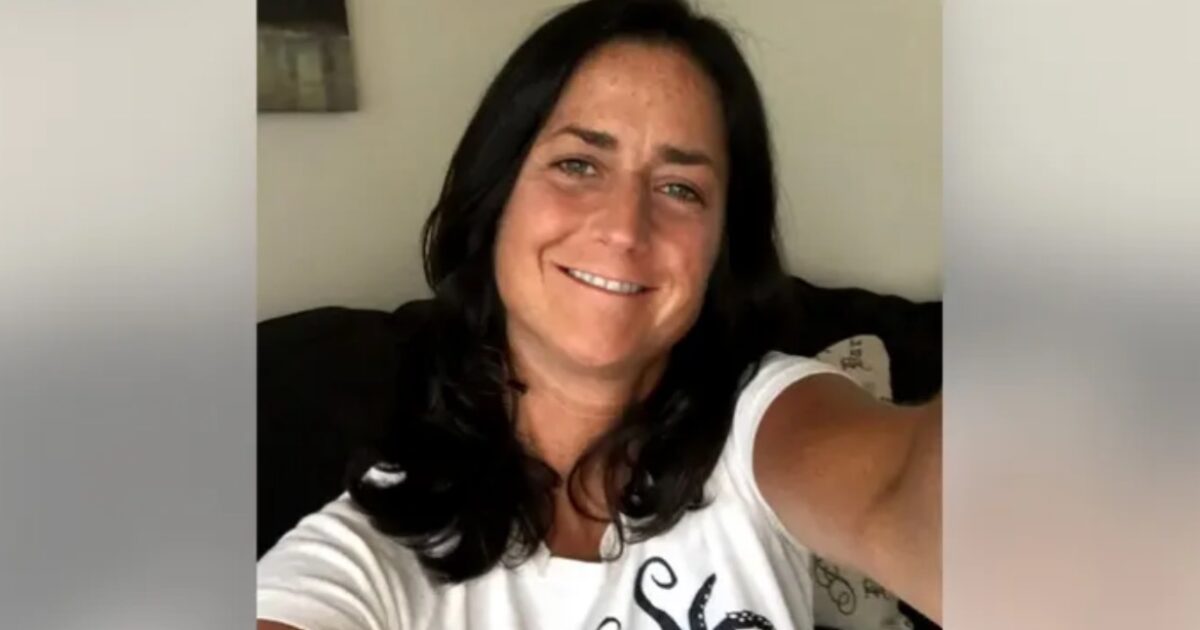 Smiling woman with long dark hair wearing a white shirt, posing for a selfie on a couch in a cozy indoor setting.