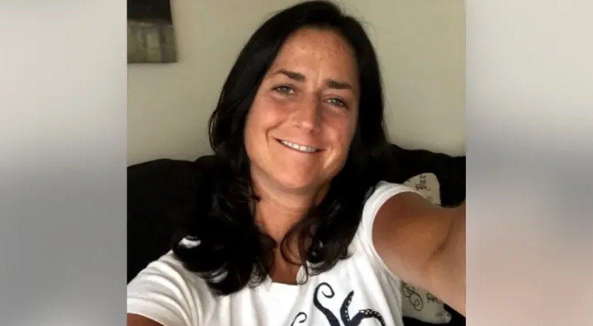 Smiling woman with long dark hair wearing a white shirt, posing for a selfie on a couch in a cozy indoor setting.