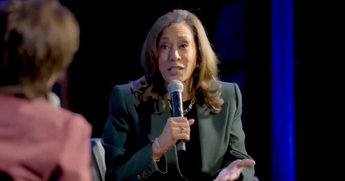 A woman in a green blazer speaks passionately into a microphone during a panel discussion, engaging with another participant off-screen.