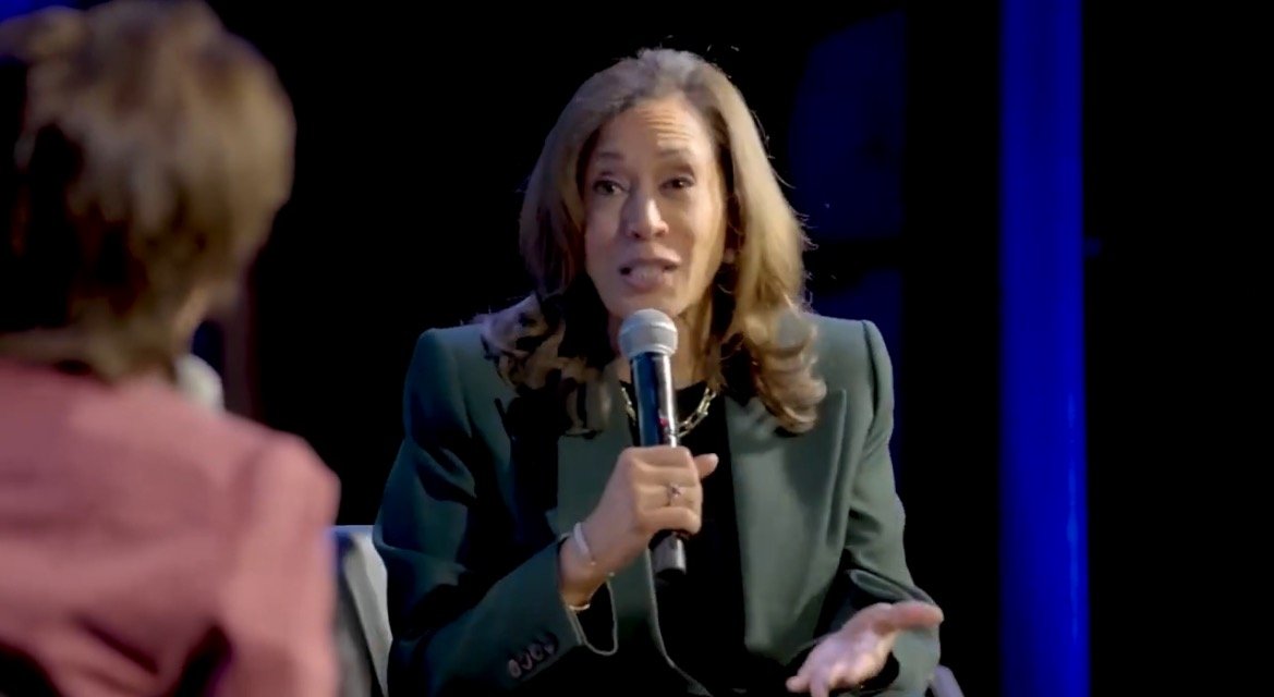 A woman in a green blazer speaks passionately into a microphone during a panel discussion, engaging with another participant off-screen.