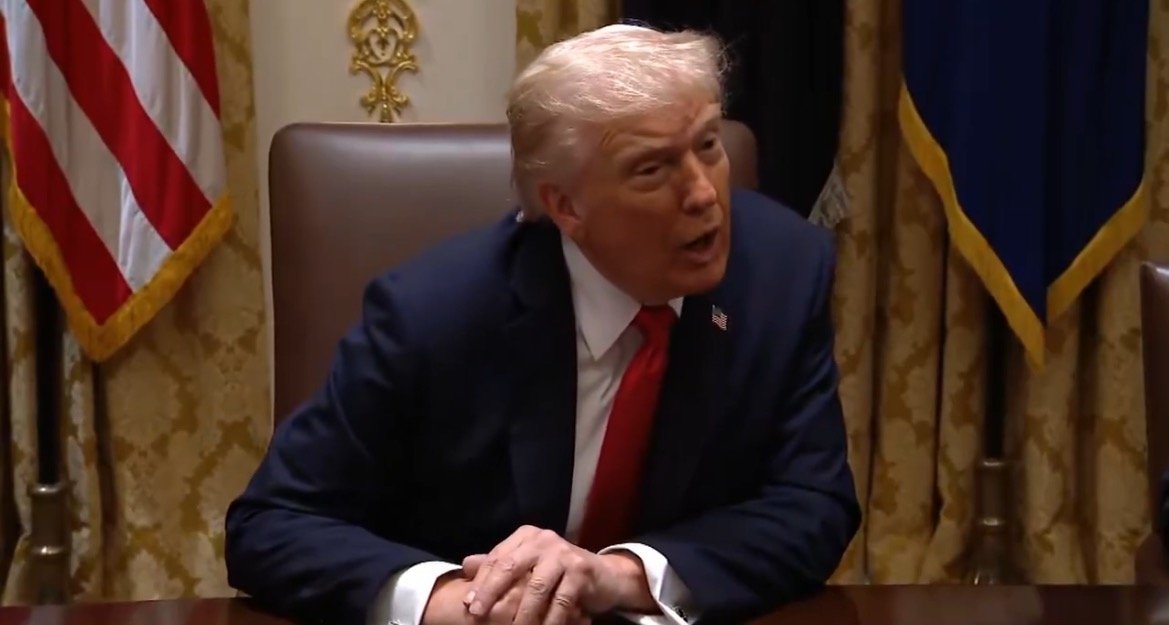 Donald Trump speaking during a meeting in the Oval Office, with American flags and drapery in the background.